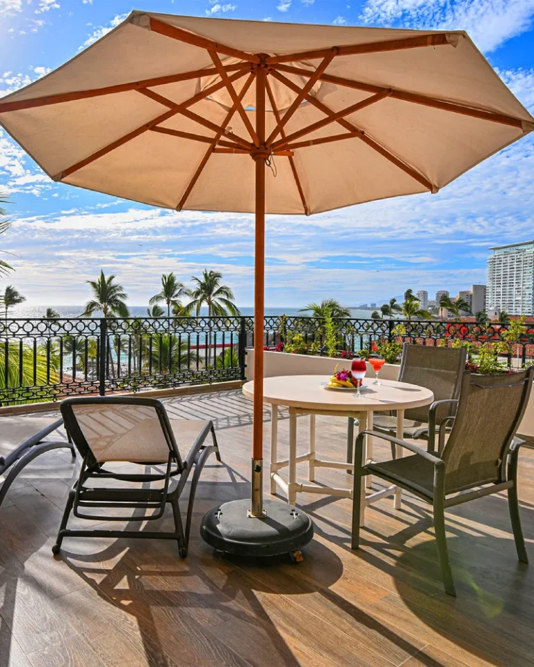 Terraza con sombrilla, mesa servida y vista al mar con palmeras.