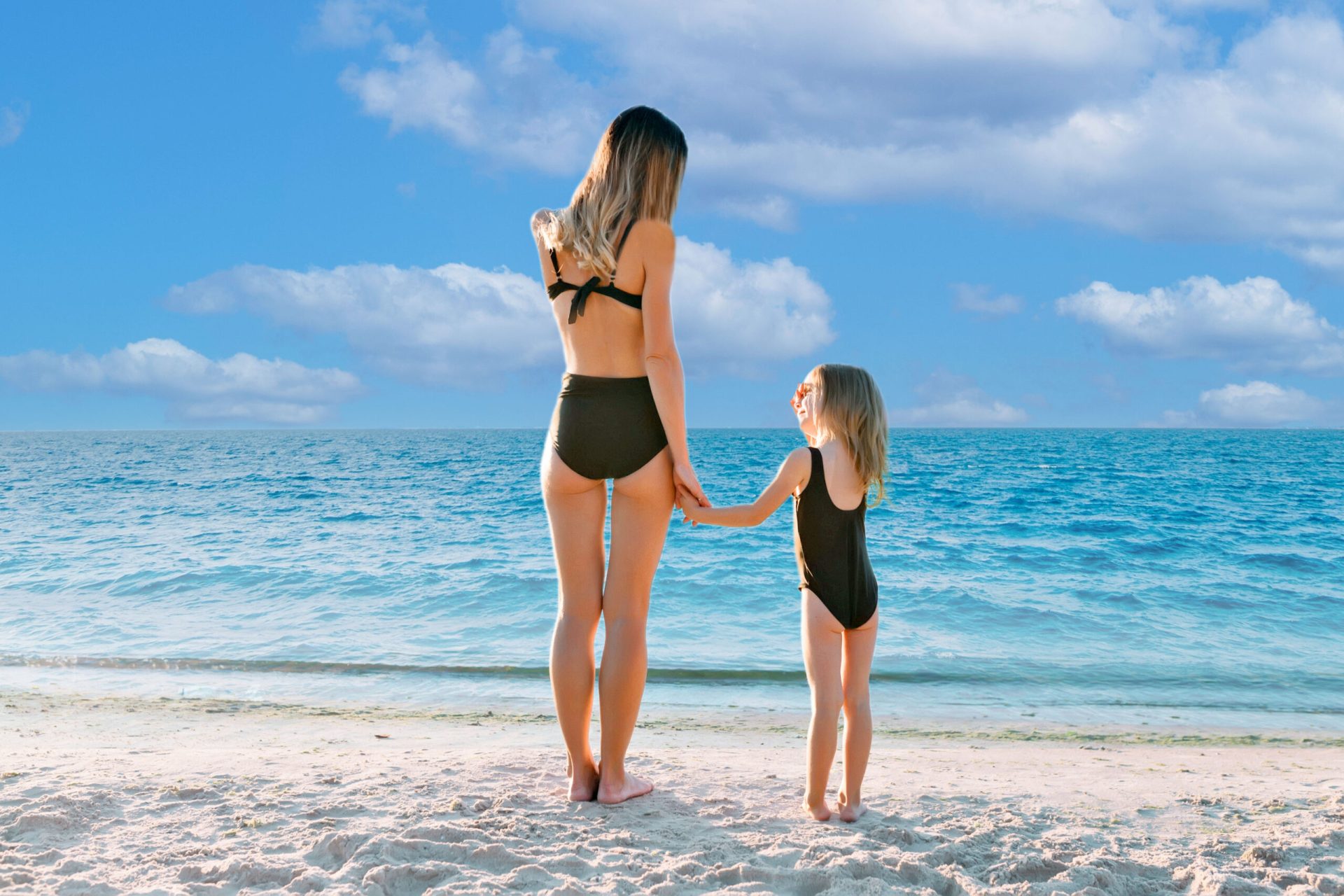 Adulto y niño de pie en la playa mirando al mar, tomados de la mano bajo un cielo parcialmente nublado, en un momento tranquilo y familiar.